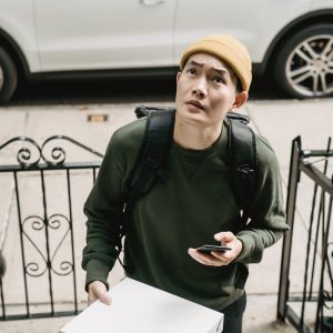 Asian man in beanie holding a pizza box and smartphone outdoors near a white car and gate.